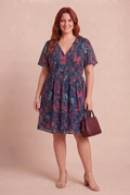 Mannequin rousse portant la robe Lavande marine avec un sac bordeaux sur fond beige rosé – mode grande taille femme